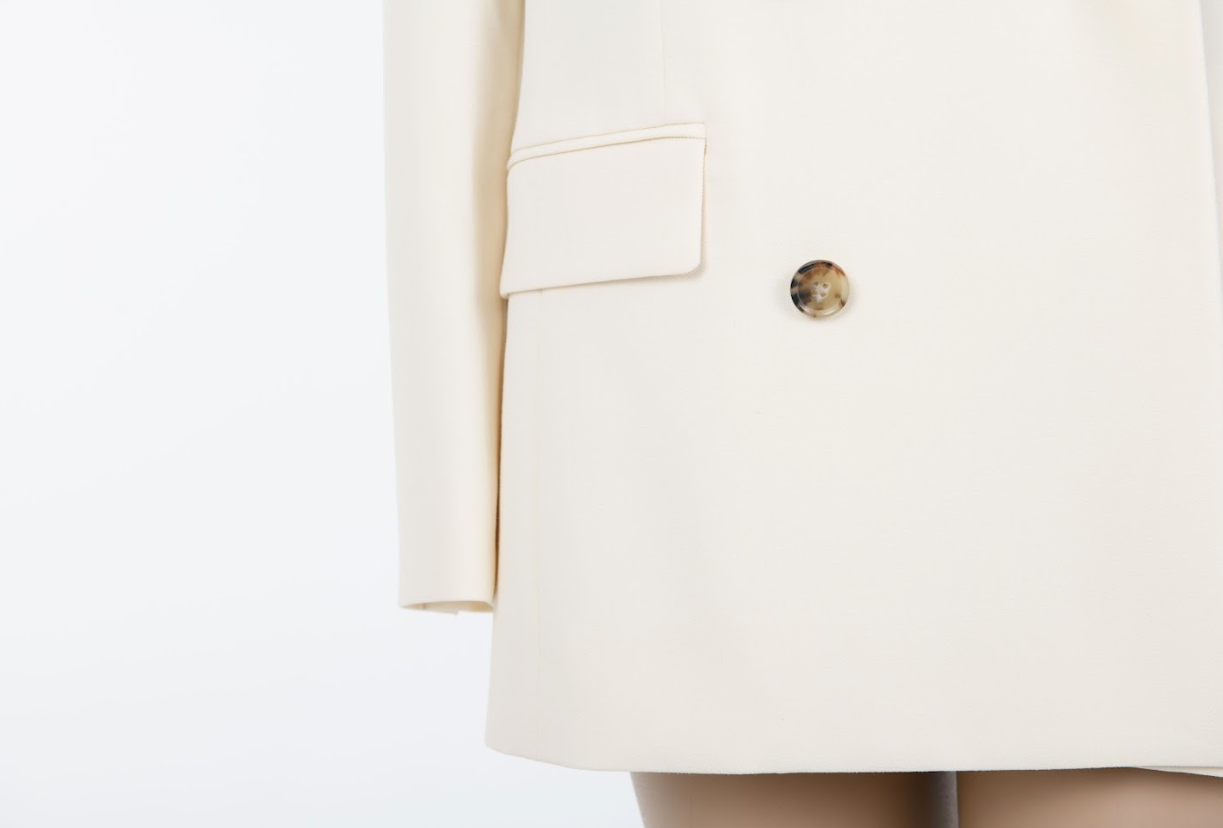 Macro detail of the FRAME Relaxed Double-Breasted Blazer showing the refined weave of the ivory fabric and the polished finish of the tortoise-style buttons.